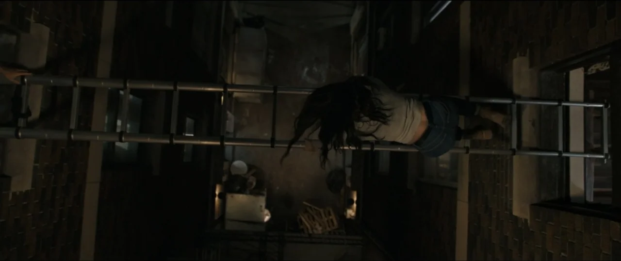 Scream VI (2023) movie still: wide — A dark urban alleyway features brick walls and a spanning metal ladder suspended high. A woman lies…