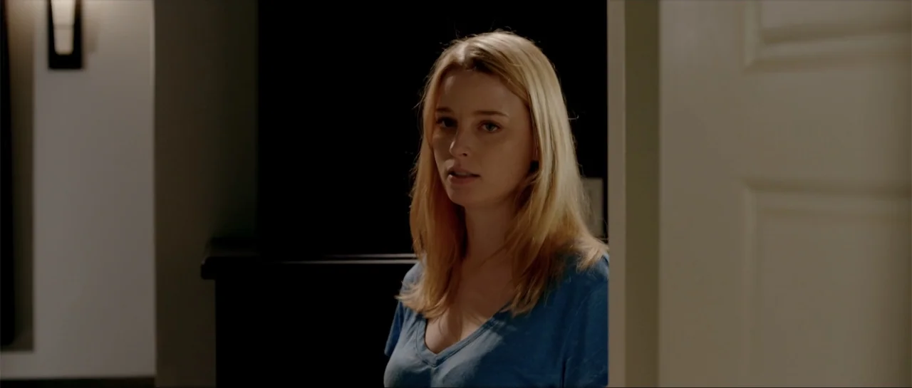 Rage (2014) movie still: medium — A woman stands in a partially lit doorway between a dark room and a hallway. She looks directly toward the camera…