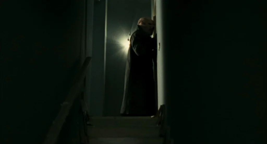 Timecrimes (2007) movie still: fullbody — A dark, narrow indoor staircase leads up toward a single light source. A person in a long coat stands at…