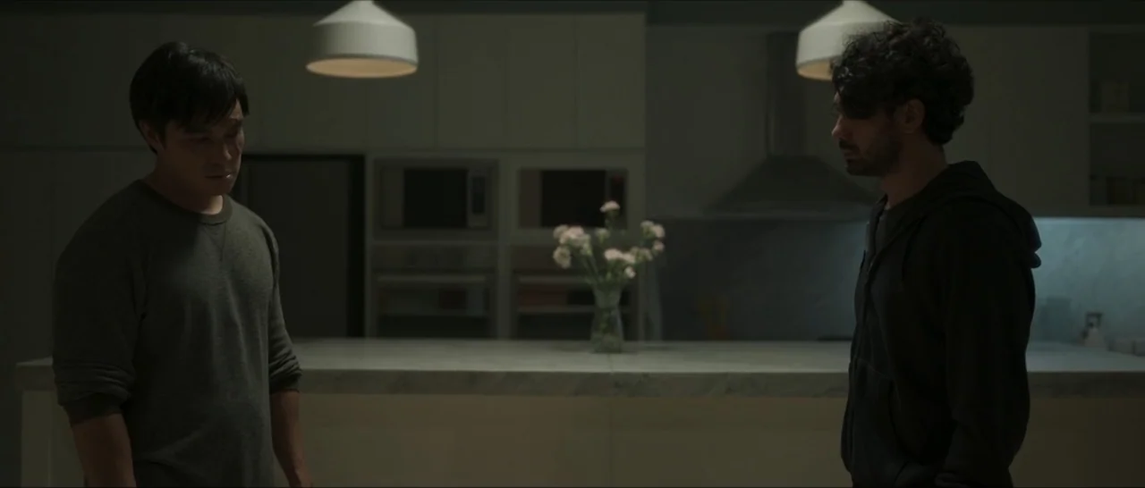 Innocent Vengeance (2023) movie still: medium — A modern kitchen island with a marble countertop holds a small vase of flowers. Two men stand on…