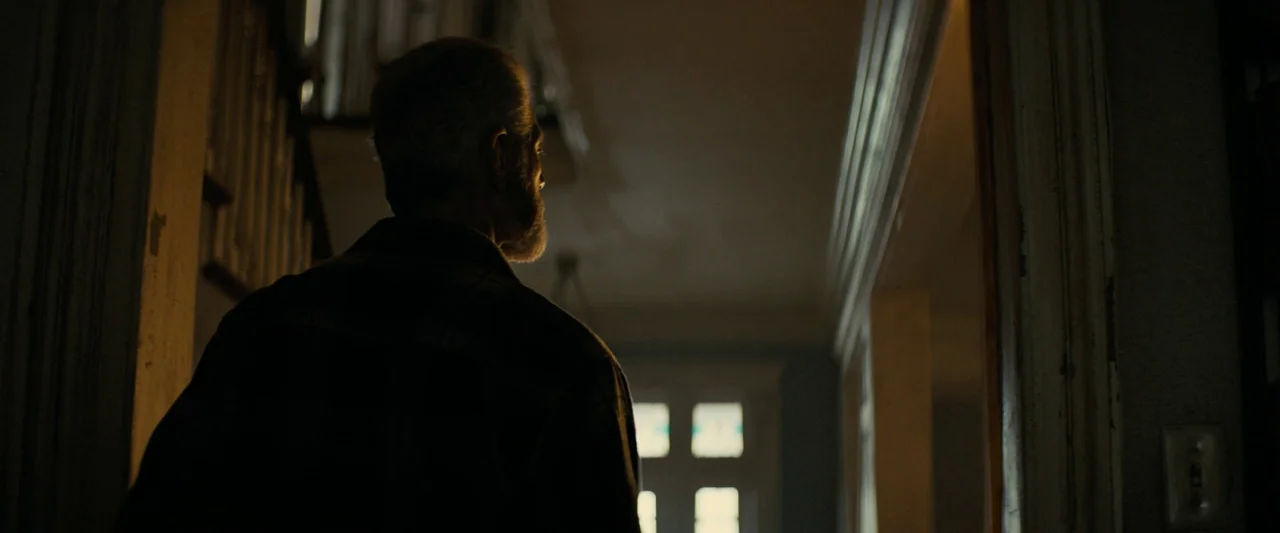 Don’t Breathe 2 (2021) movie still: medium — A dimly lit indoor residential hallway leads toward a bright doorway with glass panes. An older man…