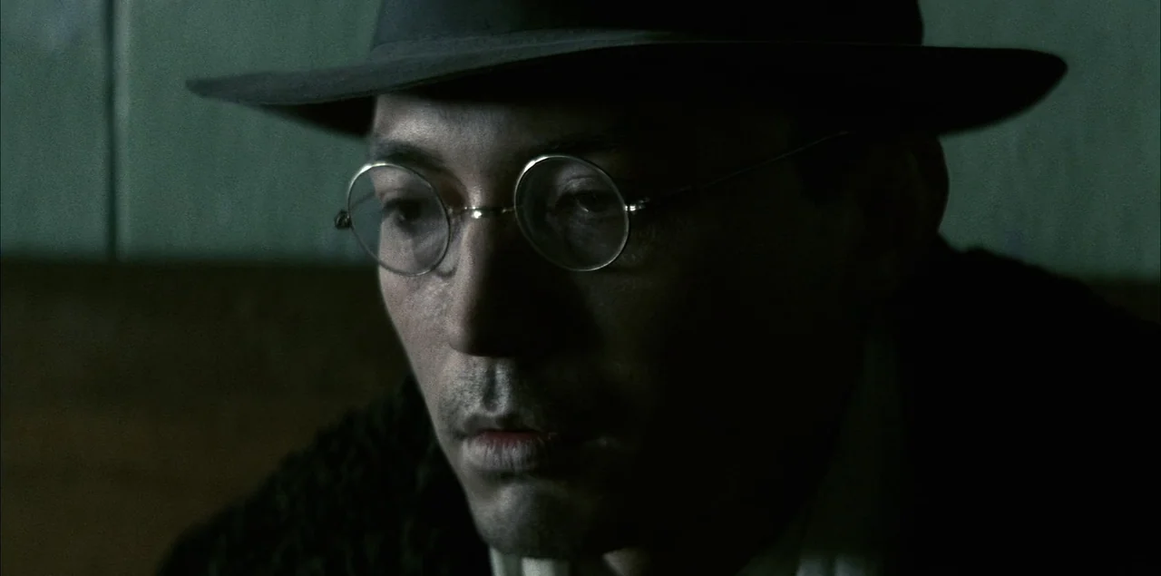The Last Emperor (1987) movie still: closeup — A dark space with indistinct walls serves as the backdrop for the subject. A man wearing a fedora and…