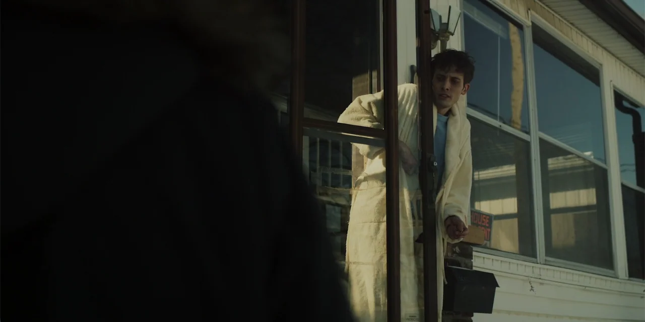 Lobos (2019) movie still: medium — A man stands in the doorway of a house on a residential street. He wears a bathrobe and peers outward at someone…
