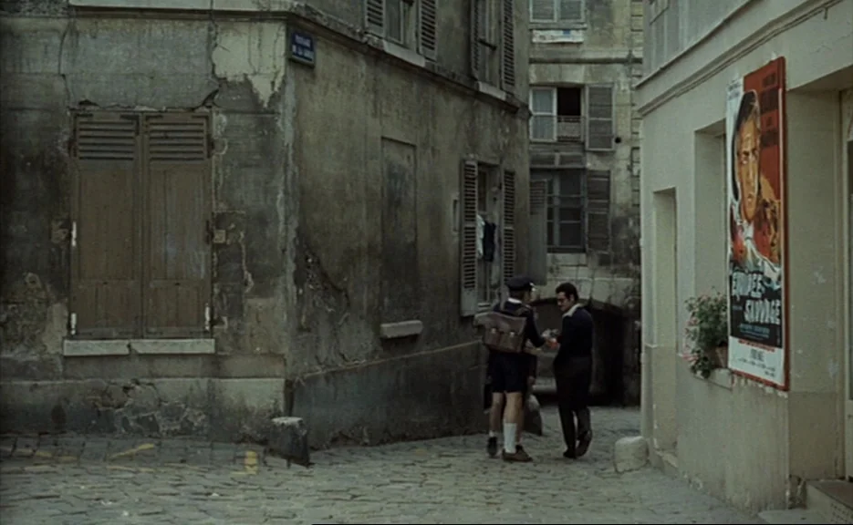 Murmur of the Heart (1971) movie still: wide — A narrow cobblestone alley flanked by weathered stone buildings with closed shutters. Two men stand…