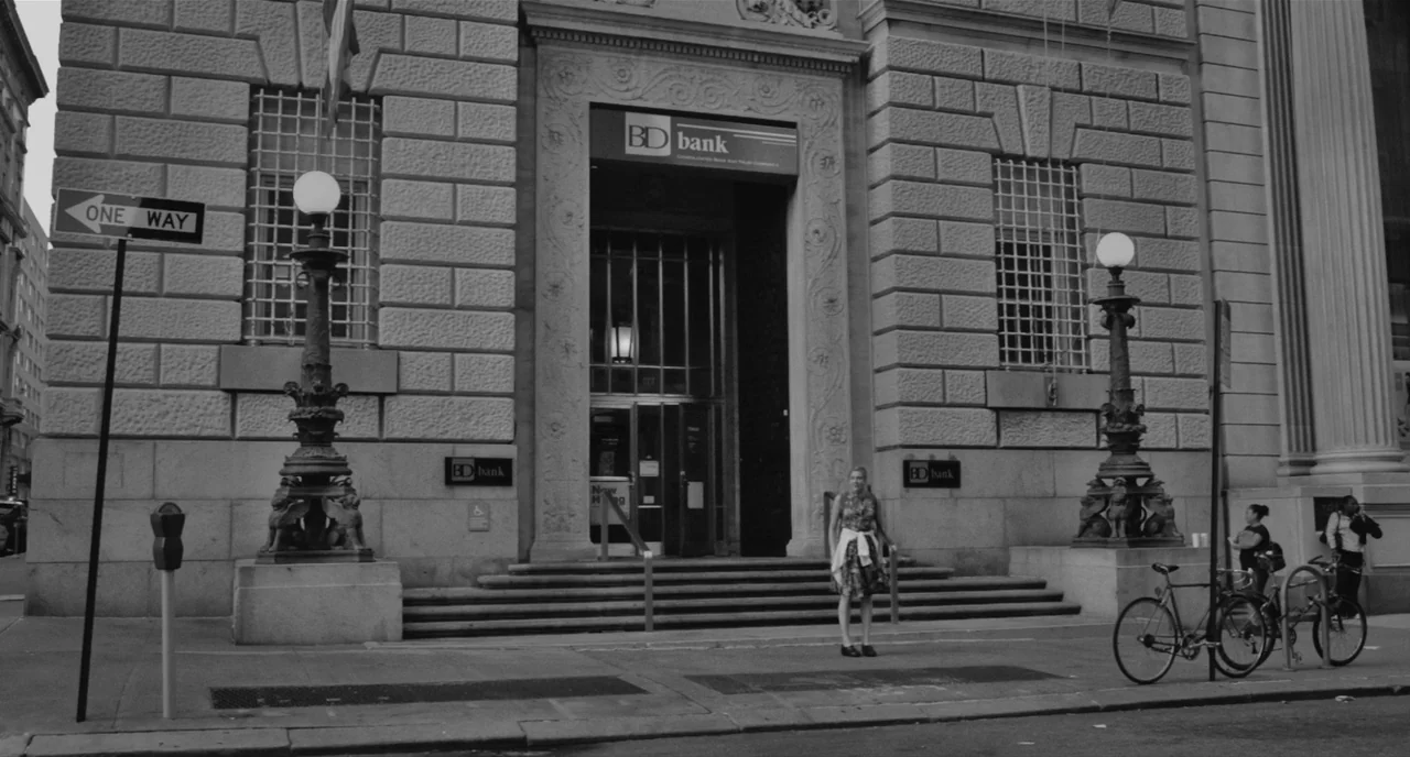 Frances Ha (2012) movie still: wide — A stone bank building features ornate carved door frames and two ornate lamp posts. A young woman stands on…