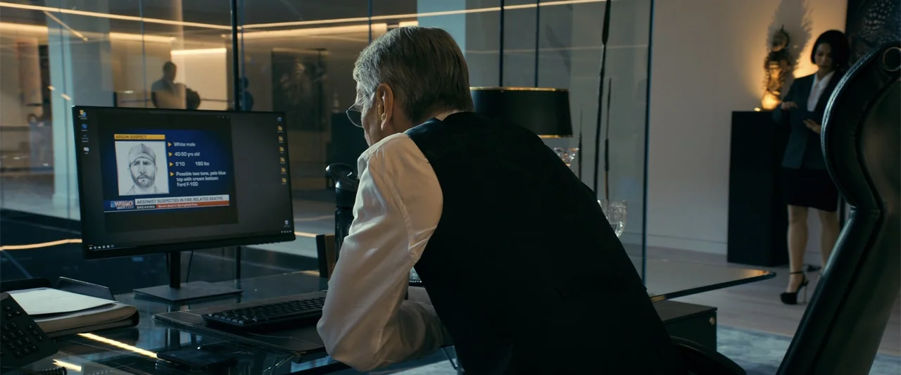 The Beekeeper (2024) movie still: medium — A modern office environment with glass partitions and a sleek glass desk. A man observes an arson suspect…