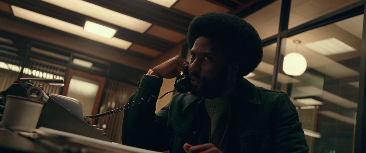 BlacKkKlansman (2018) movie still: medium — A 1970s workplace interior features wood-paneled accents and glass partitions under fluorescent ceiling…