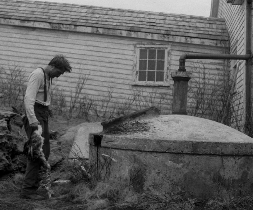 The Lighthouse (2019) movie still: fullbody — A man stands beside a concrete cistern against a white-sided building with wooden shingles. He holds a…