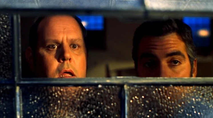 Out Of Sight (1998) movie still: closeup — Two men look through a textured glass windowpane inside a dimly lit area. They are staring ahead with…