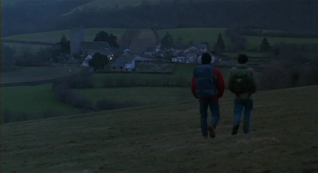 An American Werewolf in London (1981) movie still: wide — Two hikers walk down a grassy slope overlooking a rural English village at twilight. The…