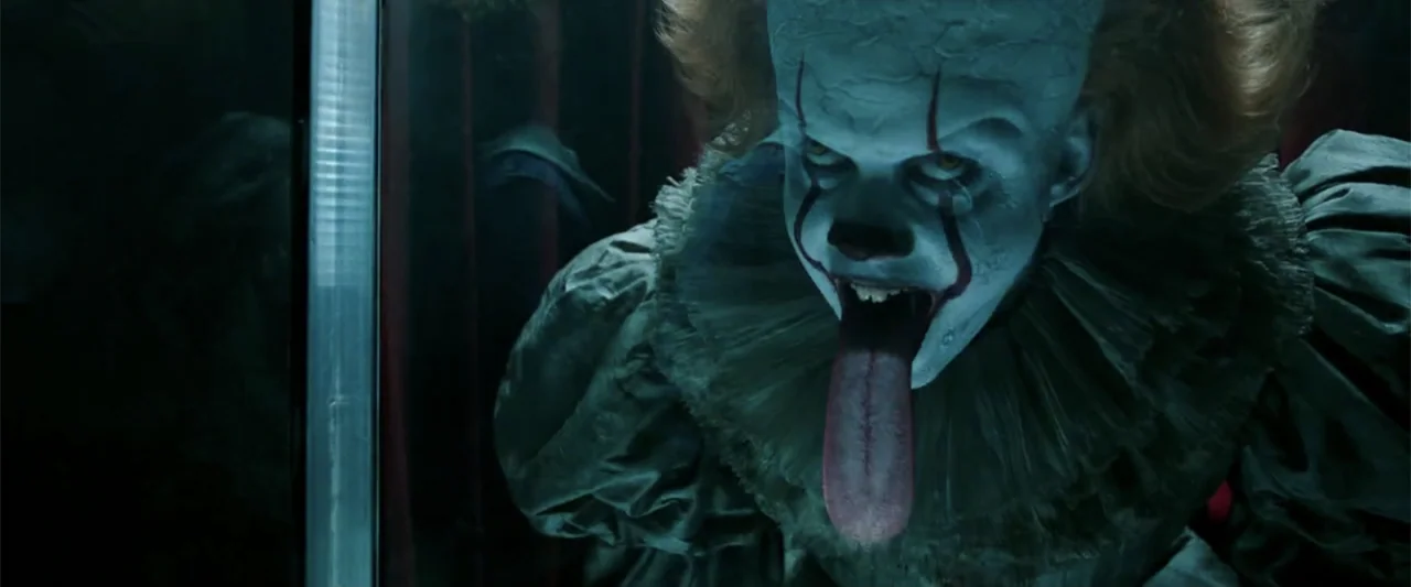 K.G.F: Chapter 2 (2022) movie still: closeup — A dark indoor setting features reflective glass partitions framing a pale clown figure. The costumed…