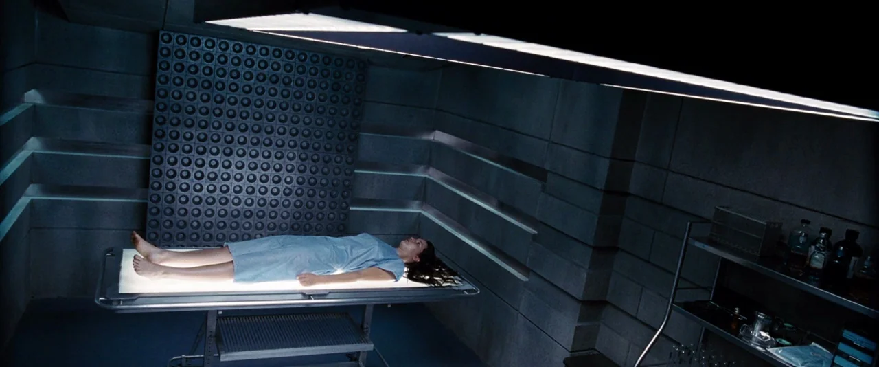 The Hunger Games (2012) movie still: wide — A clinical, windowless room features concrete-block walls with a distinctive geometric grid pattern. A…