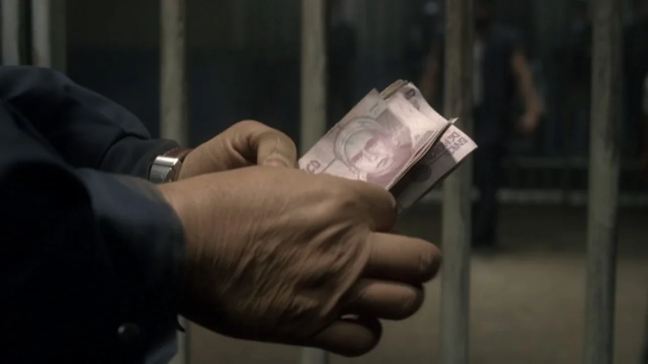 The Shepherd (2023) movie still: closeup — A dimly lit interior space features vertical metal bars in the background. Two hands hold a small stack…