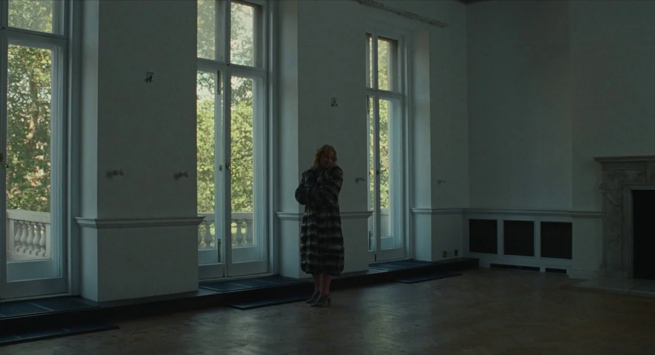 The Nest (2020) movie still: wide — A spacious, sparsely furnished drawing room features high ceilings and tall, classic glass doors. A solitary…