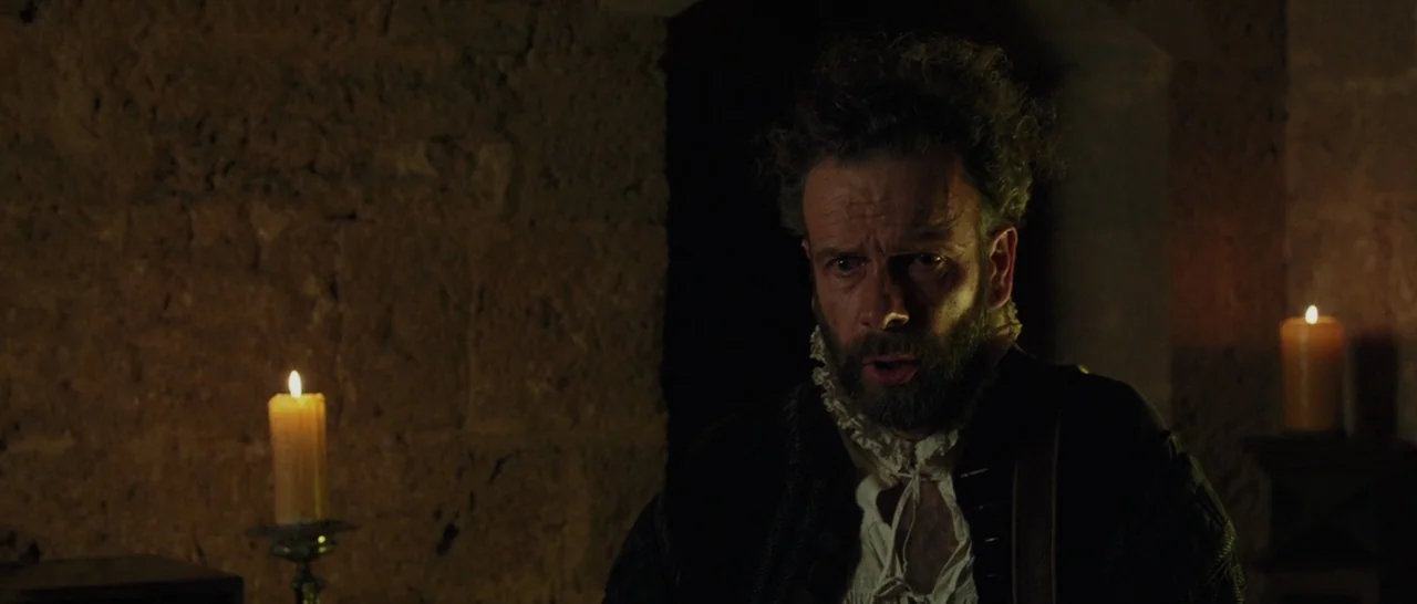 Tale of Tales (2015) movie still: medium — A dark, textured stone room illuminated by the glow of two lit candles. A bearded man in period clothing…