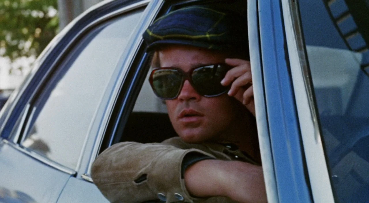 The Iguana with the Tongue of Fire (1971) movie still: closeup — A man sits in the driver seat of a vintage vehicle during daylight. He wears dark…