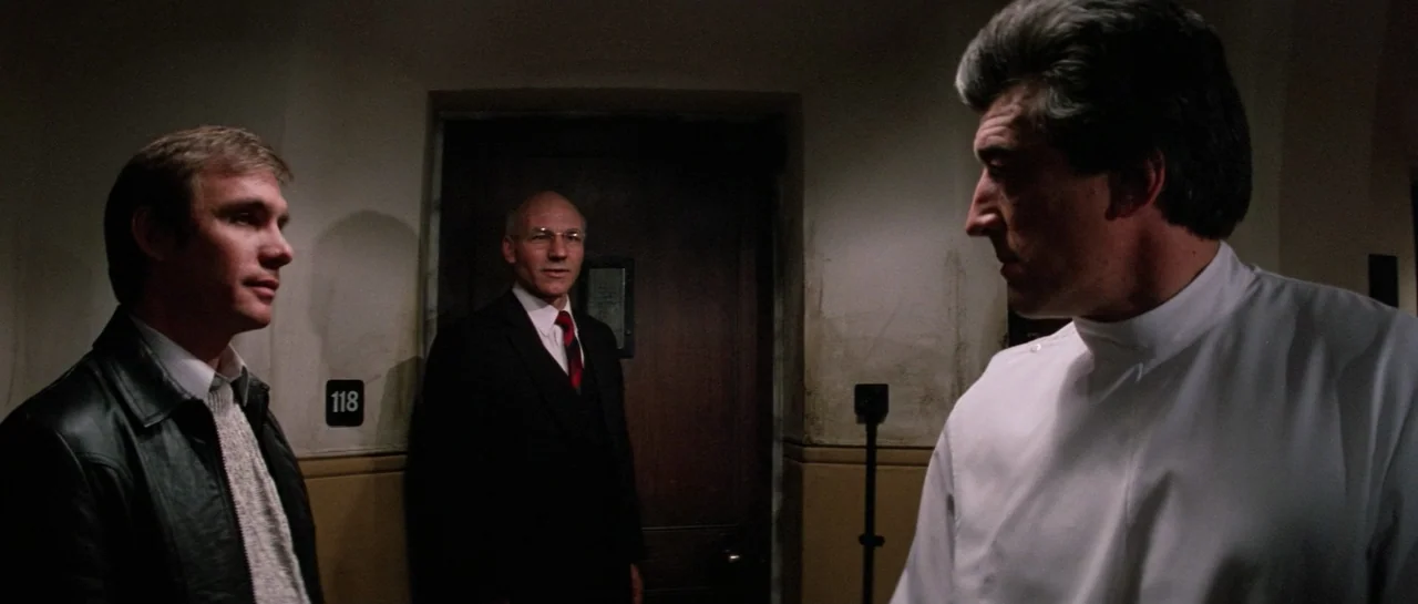 Lifeforce (1985) movie still: medium — A dimly lit hospital corridor features a wooden door labeled 118. Three men stand in the hallway while…