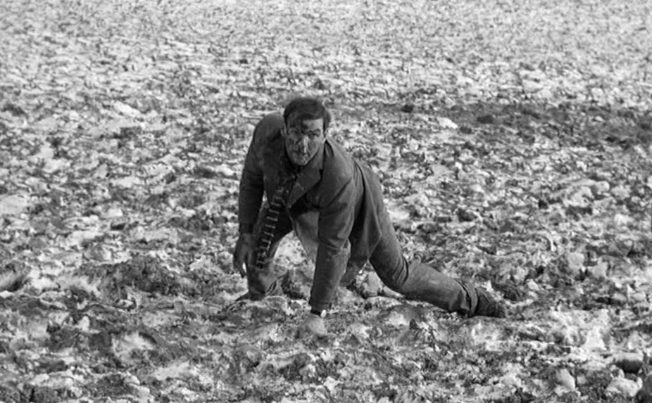 The Criminal (1960) movie still: fullbody — An uneven, churned mud field stretches across the entire frame under overcast skies. A man in a…