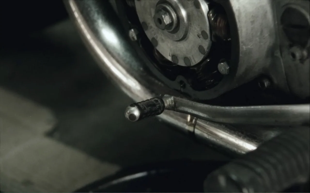 The Greasy Hands Preachers (2014) movie still: closeup — A close-up of a greasy motorcycle engine housing and kick-start lever. Metallic components…