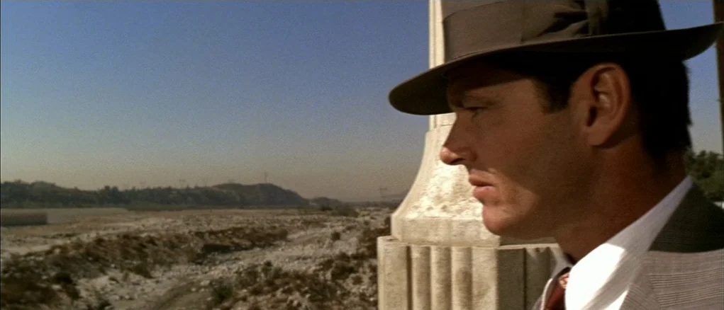 Chinatown (1974) movie still: closeup — A man in a fedora stands on a concrete bridge overlooking a dry riverbed. The landscape is sparse and dusty…