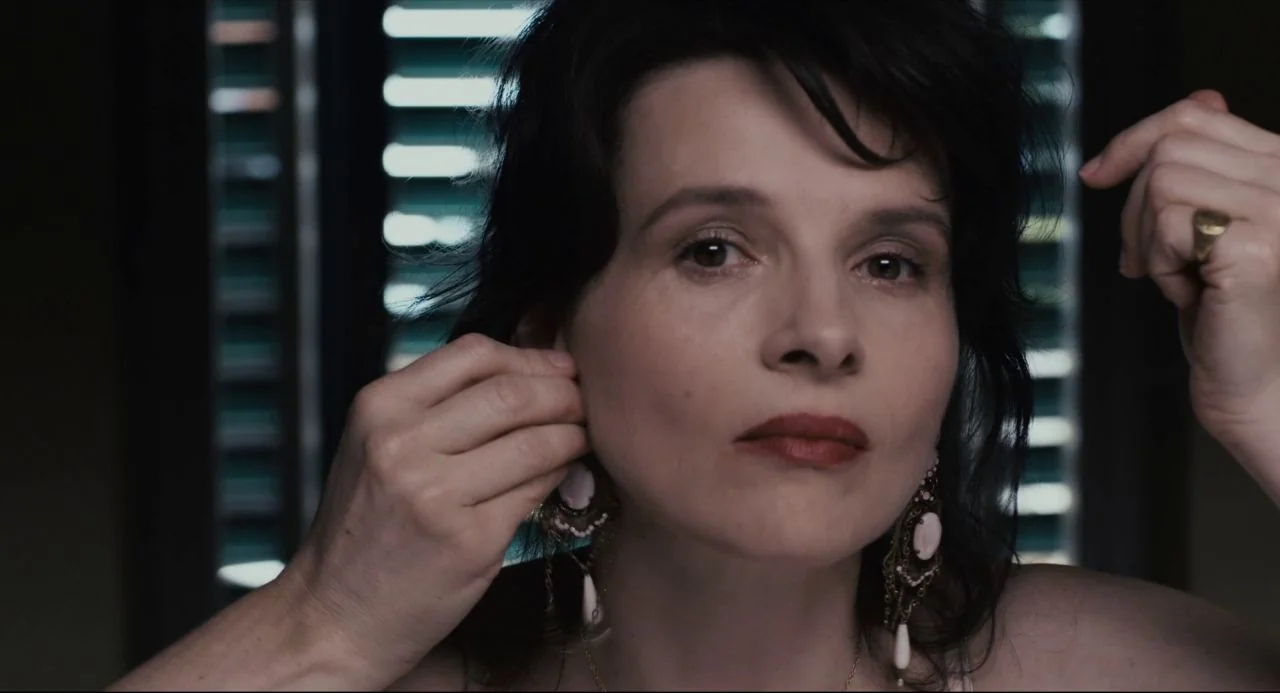 Certified Copy (2010) movie still: closeup — A woman stands before a reflective surface in a space with dark slatted blinds. She uses both hands to…