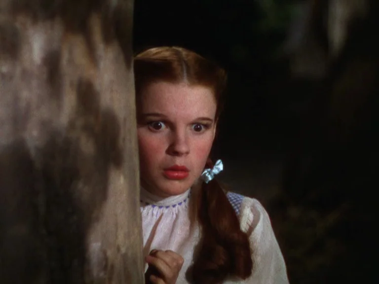 The Wizard Of Oz (1939) movie still: closeup — A young girl with pigtails peeks from behind the rough bark of a tree. The dark shadows of the…