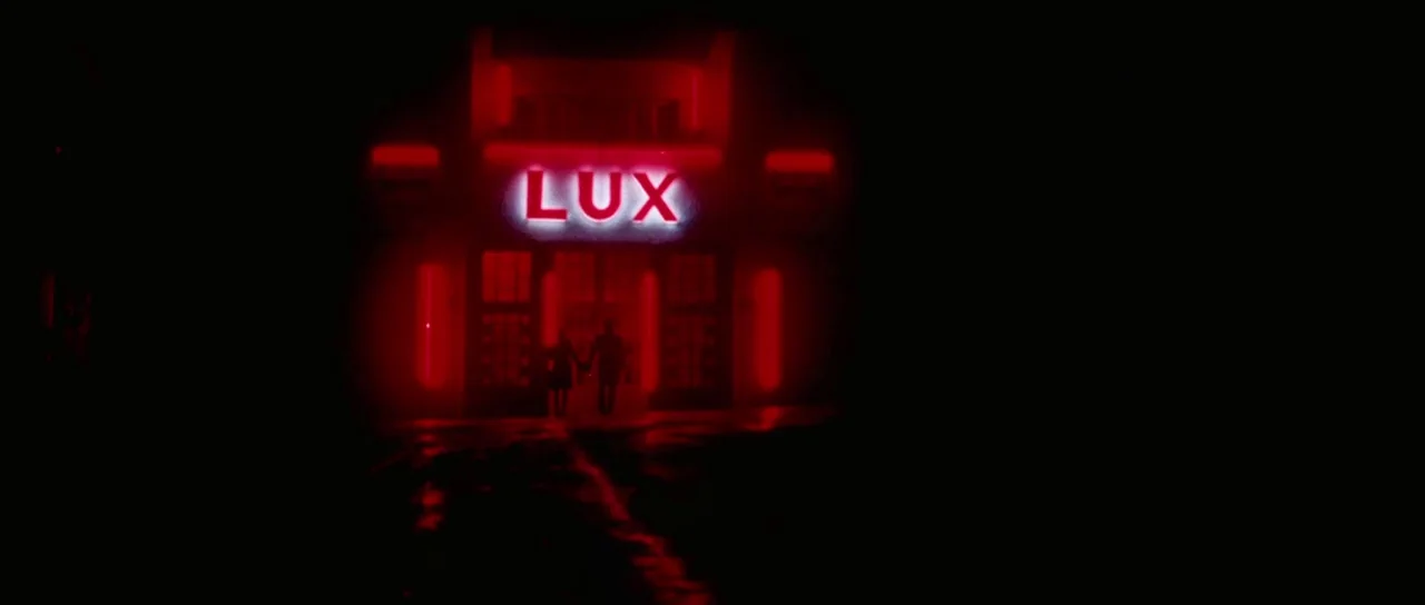 Alléluia (2014) movie still: wide — The exterior of a building features a bright red neon sign reading LUX above. A man and woman hold hands while…