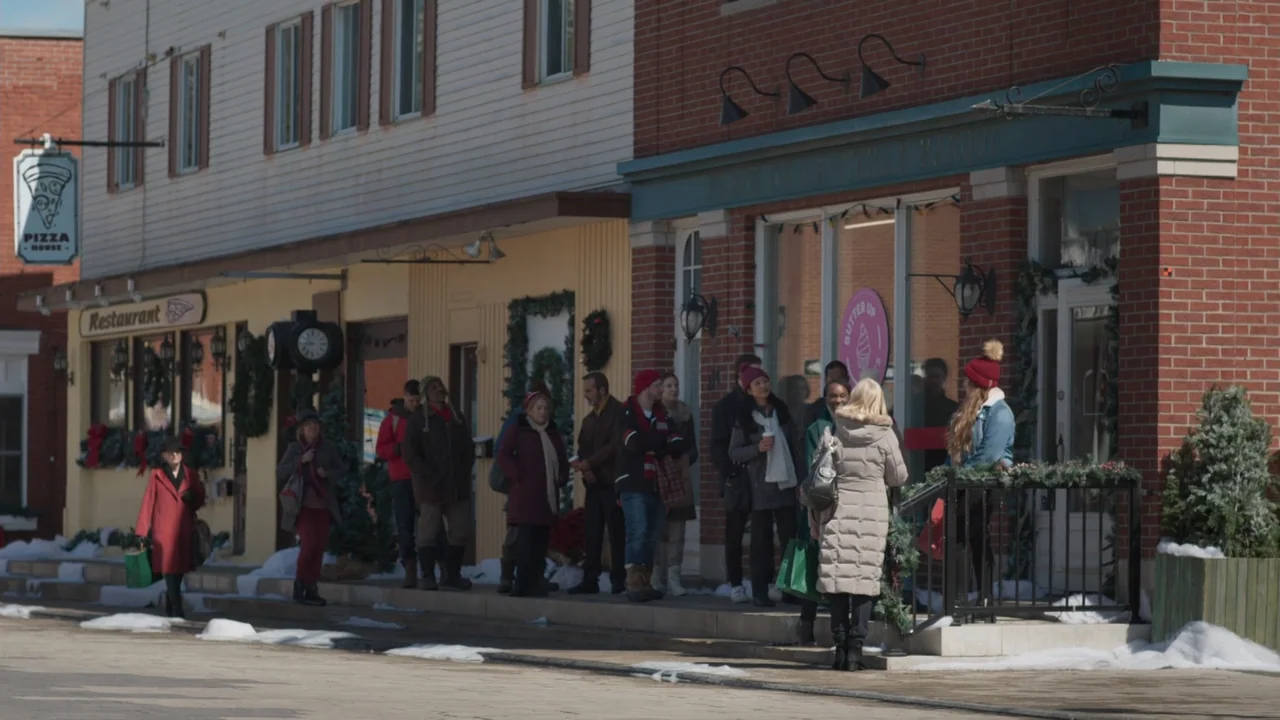 A Gingerbread Christmas (2022) movie still: wide — A row of brick and painted storefronts on a sidewalk features Christmas decorations. A group of…