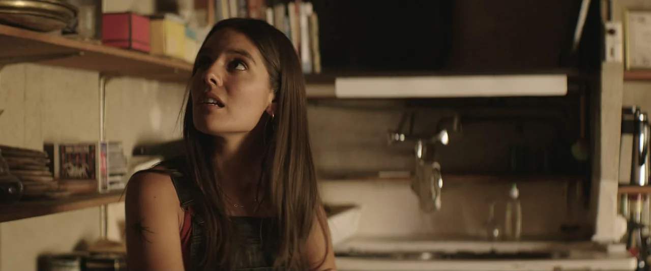 The Only One (2021) movie still: medium — A cluttered space with wooden shelves holding books and various objects in the background. A woman in…