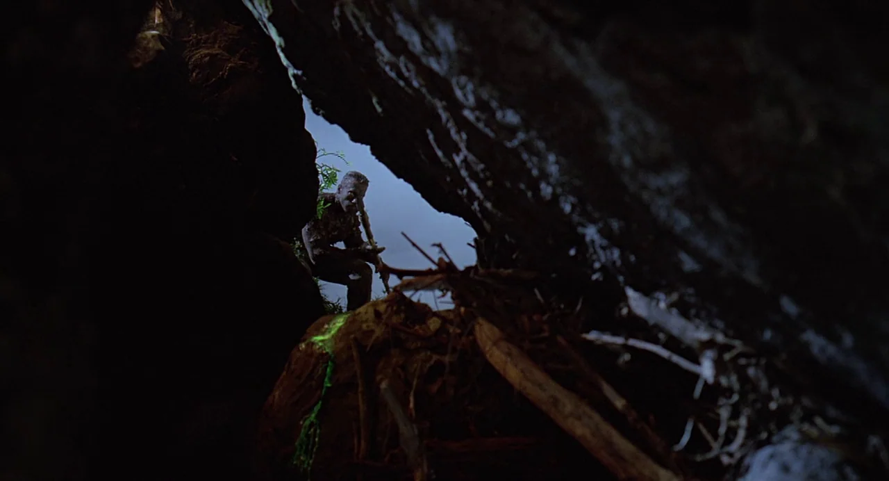 Predator (1987) movie still: wide — A dark, jagged rock formation creates a natural frame around an opening to daylight. A person in camouflage gear…