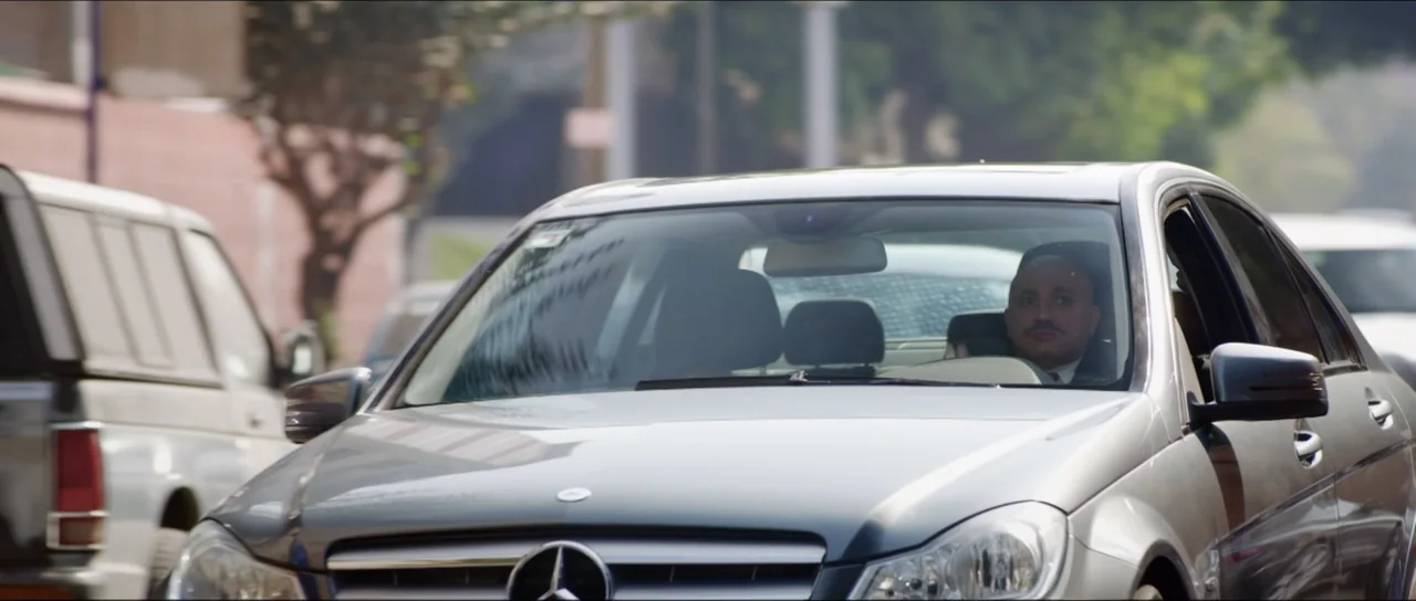 Mirreyes vs Godínez (2019) movie still: medium — A silver sedan drives down a paved urban road during the daytime. A man with a mustache sits in the…