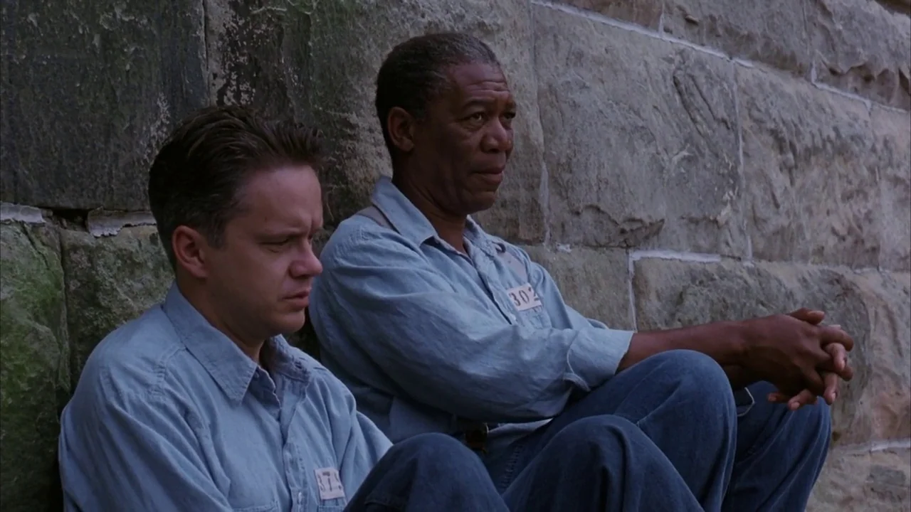 The Shawshank Redemption (1994) movie still: medium — Two men sit against the rough, textured surface of a large grey stone wall. The men wear…