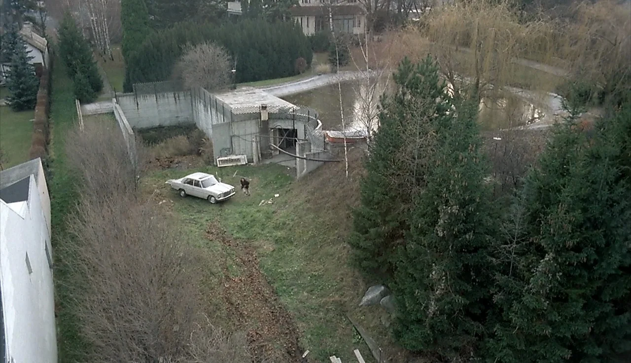Angst (1983) movie still: wide — A neglected park area features a concrete bunker structure and a small stagnant pond. A lone man walks across the…