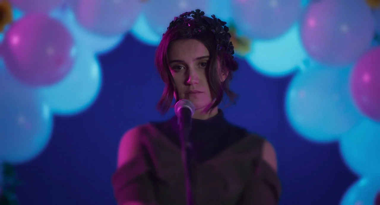 All You Need Is Death (2023) movie still: medium — A dimly lit stage features a person standing behind a microphone stand. Clusters of blurred…