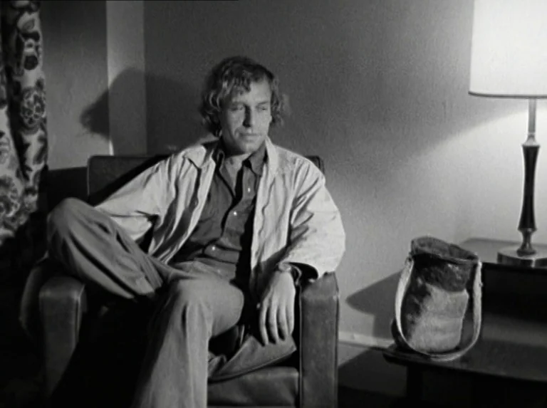 Alice in the Cities (1974) movie still: medium — A spare motel interior features a single armchair beside a lamp on a table. A man with curly hair…