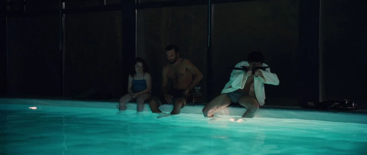 Our Day Will Come (2010) movie still: wide — A modern indoor swimming pool features floor-to-ceiling windows at night. Two men and a young girl sit…