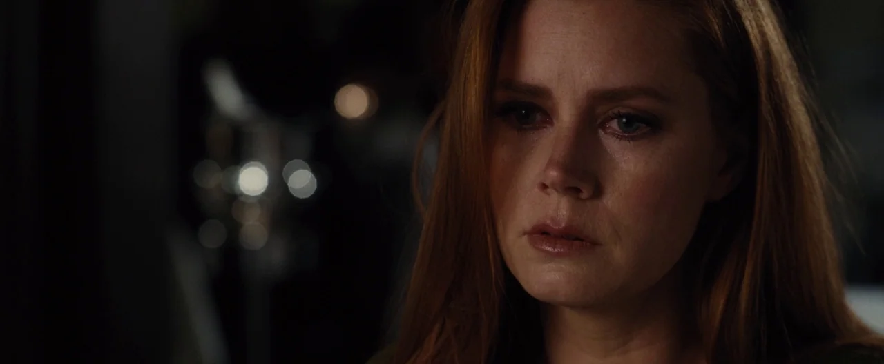 Nocturnal Animals (2016) movie still: closeup — A dark, dimly lit indoor space with blurred circular highlights in the background. A woman with long…