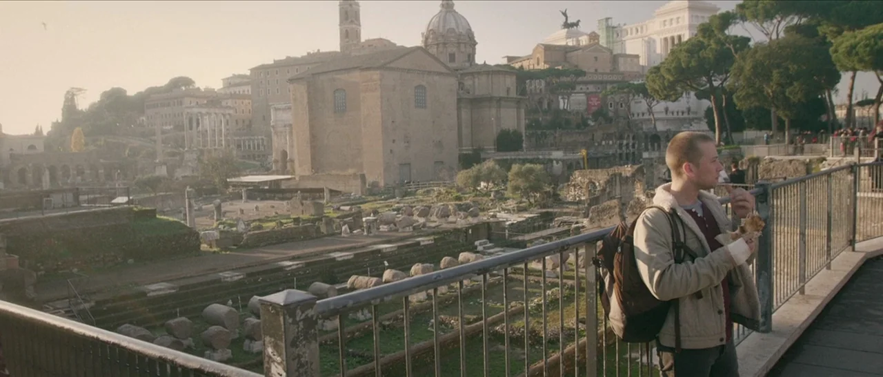 Spring (2014) movie still: medium — An overlook provides a wide perspective of the ancient ruins of the Roman Forum. A young man stands by a metal…