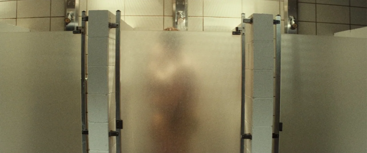 Terrifier 3 (2024) movie still: medium — A public shower area features white square wall tiles and frosted glass dividers. A person stands behind…