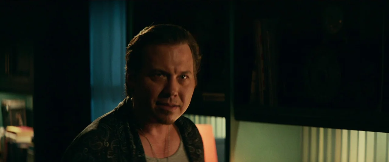 Halloween Kills (2021) movie still: medium — A dimly lit room with blurred shelves and a window in the background. A man stands in the foreground,…