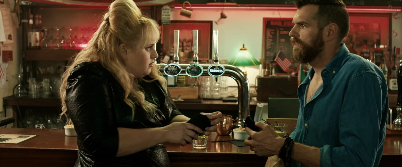 The Hustle (2019) movie still: medium — A wooden bar interior features tap handles and backlit shelving with bottles. A blonde woman and a bearded…
