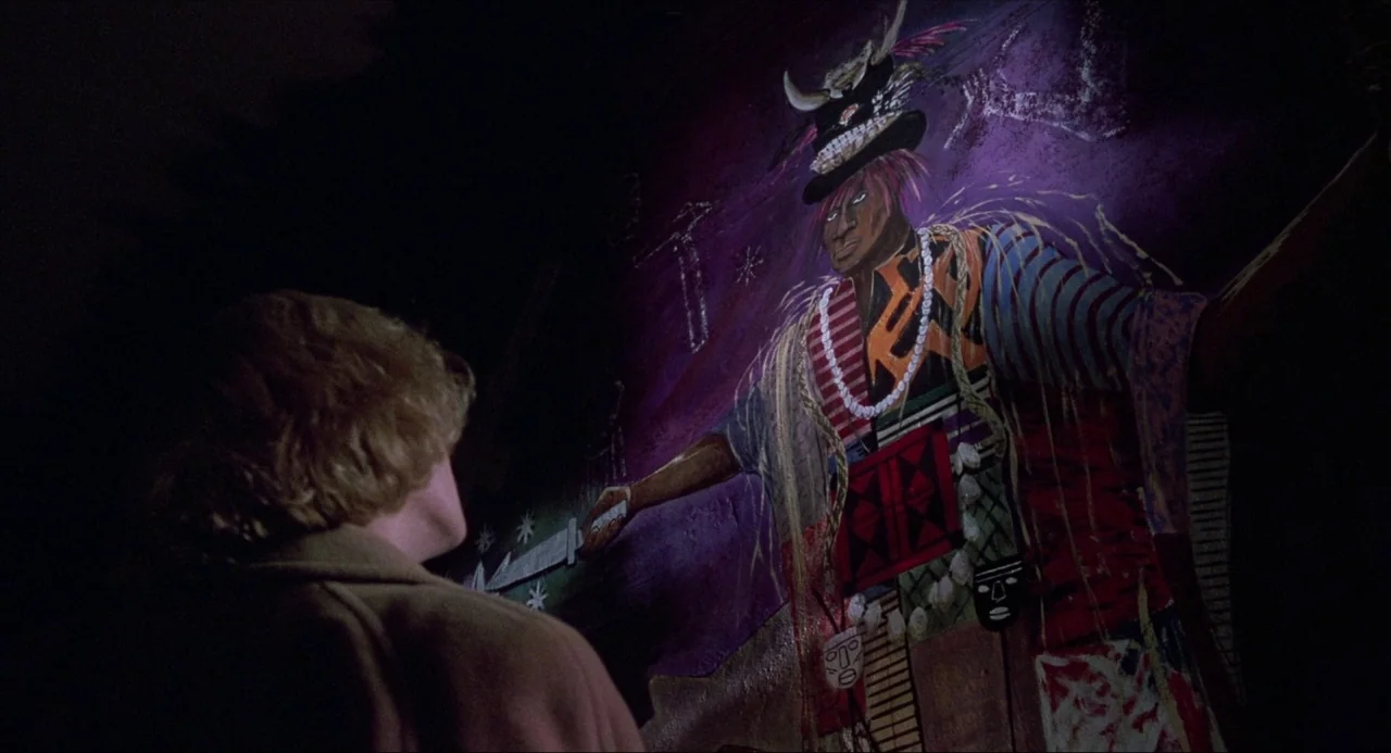 Child’s Play (1988) movie still: medium — A dark, dimly lit interior room features a prominent, brightly colored wall mural. A man stands facing the…
