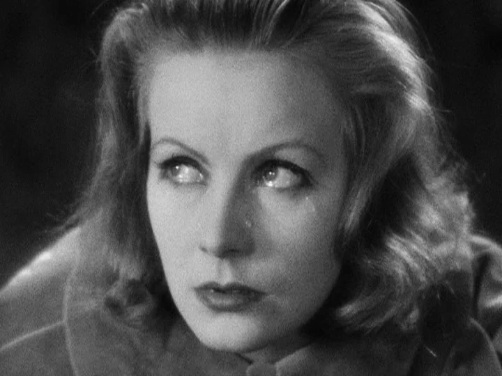 Queen Christina (1933) movie still: closeup — A dark, nondescript interior space surrounds the subject. A woman stares into the distance with a…