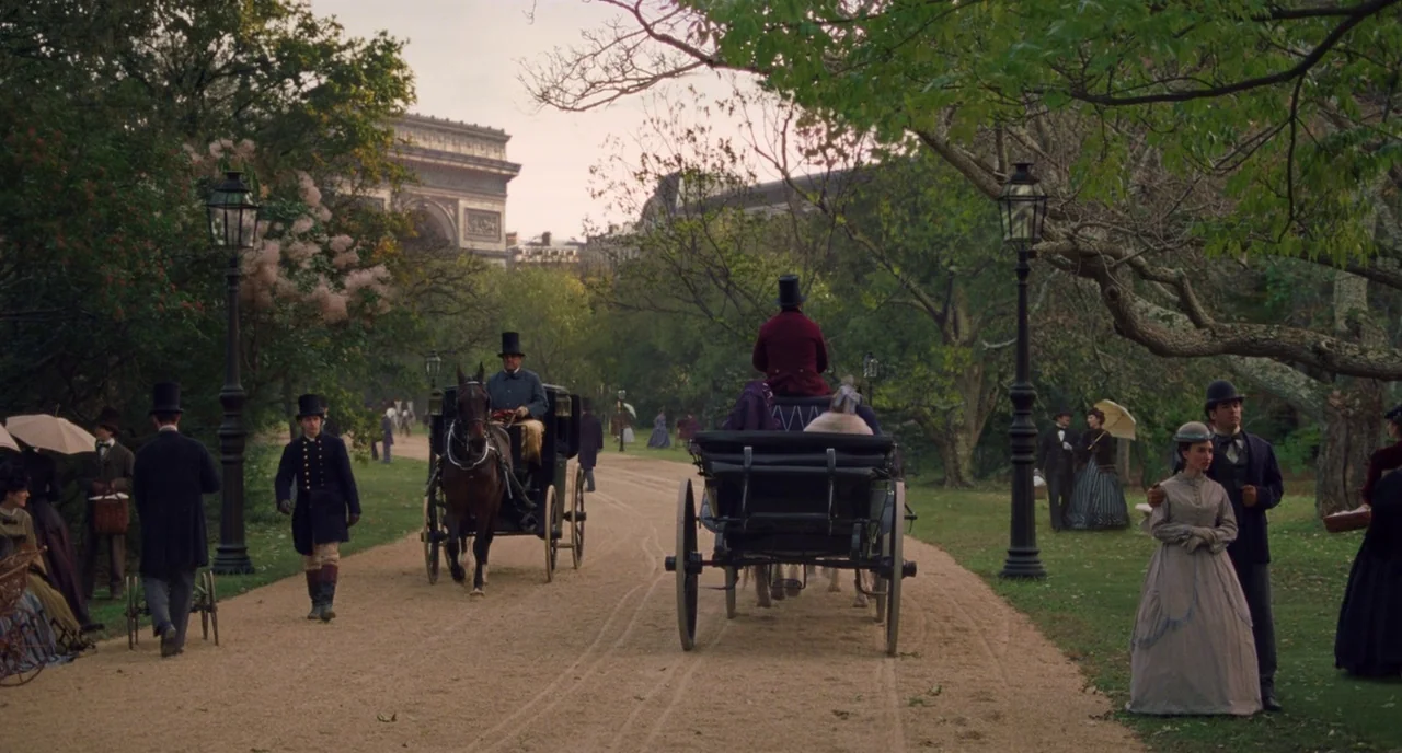 Little Women (2019) movie still: wide — A gravel pathway in a wooded park with the Arc de Triomphe visible beyond. Several figures in 19th-century…