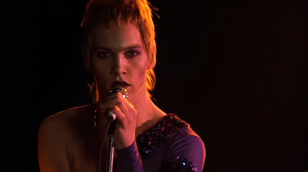 Velvet Goldmine (1998) movie still: medium — A dark environment with a single subject centered on a performance space. The individual sings into a…