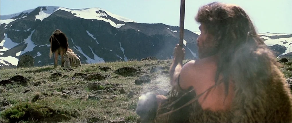 Clan of the Cave Bear (1986) movie still: medium — A high-altitude grassy mountain slope features patches of snow on the distant peaks. A man in…