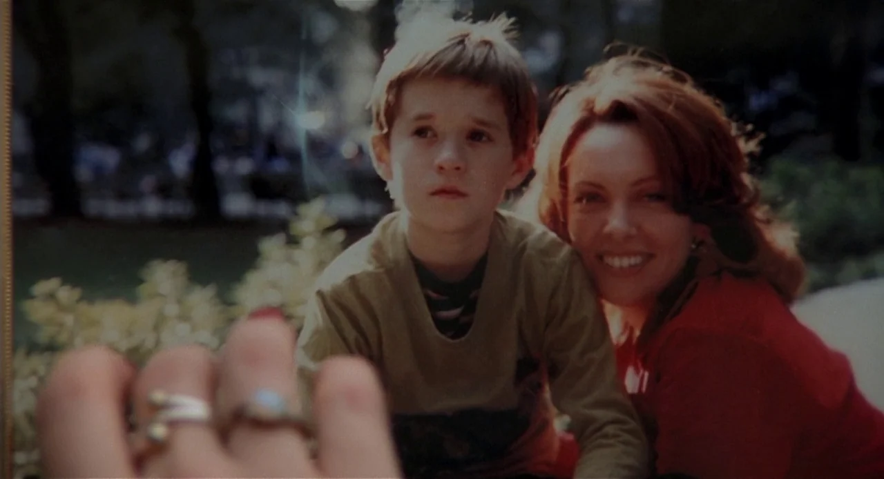 The Sixth Sense (1999) movie still: medium — A close-up photograph shows a young boy sitting next to a smiling woman. They are positioned outdoors…