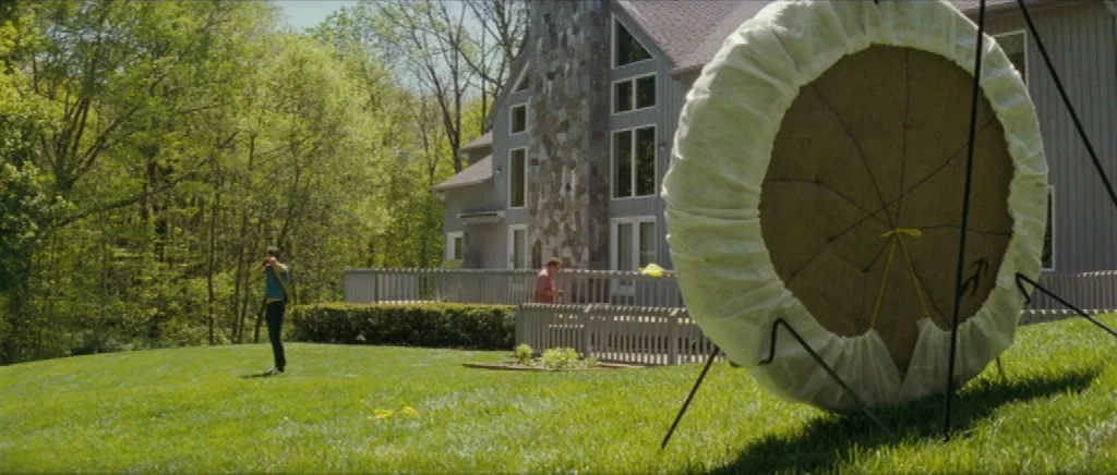 We Need To Talk About Kevin (2011) movie still: wide — A grassy lawn with a large circular archery target on a metal stand. A man stands in the…