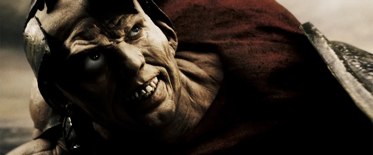 300 (2007) movie still: closeup — A scorched, war-torn exterior with a gritty, earthy atmosphere. A disfigured fighter in rustic armor glares with a…