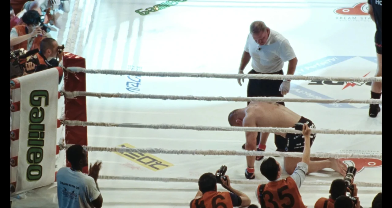 The Smashing Machine (2025) movie still: wide — A brightly lit MMA ring with a white canvas floor features multiple camera operators around it. A…